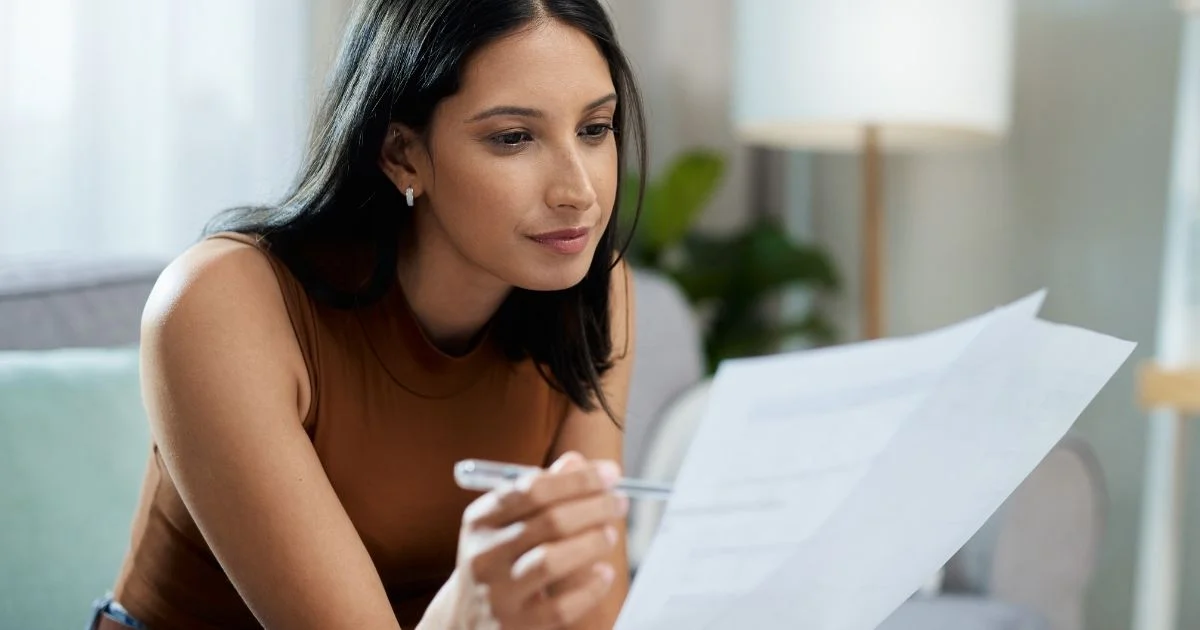 Woman sitting indoors reading paperwork with pen in hand.